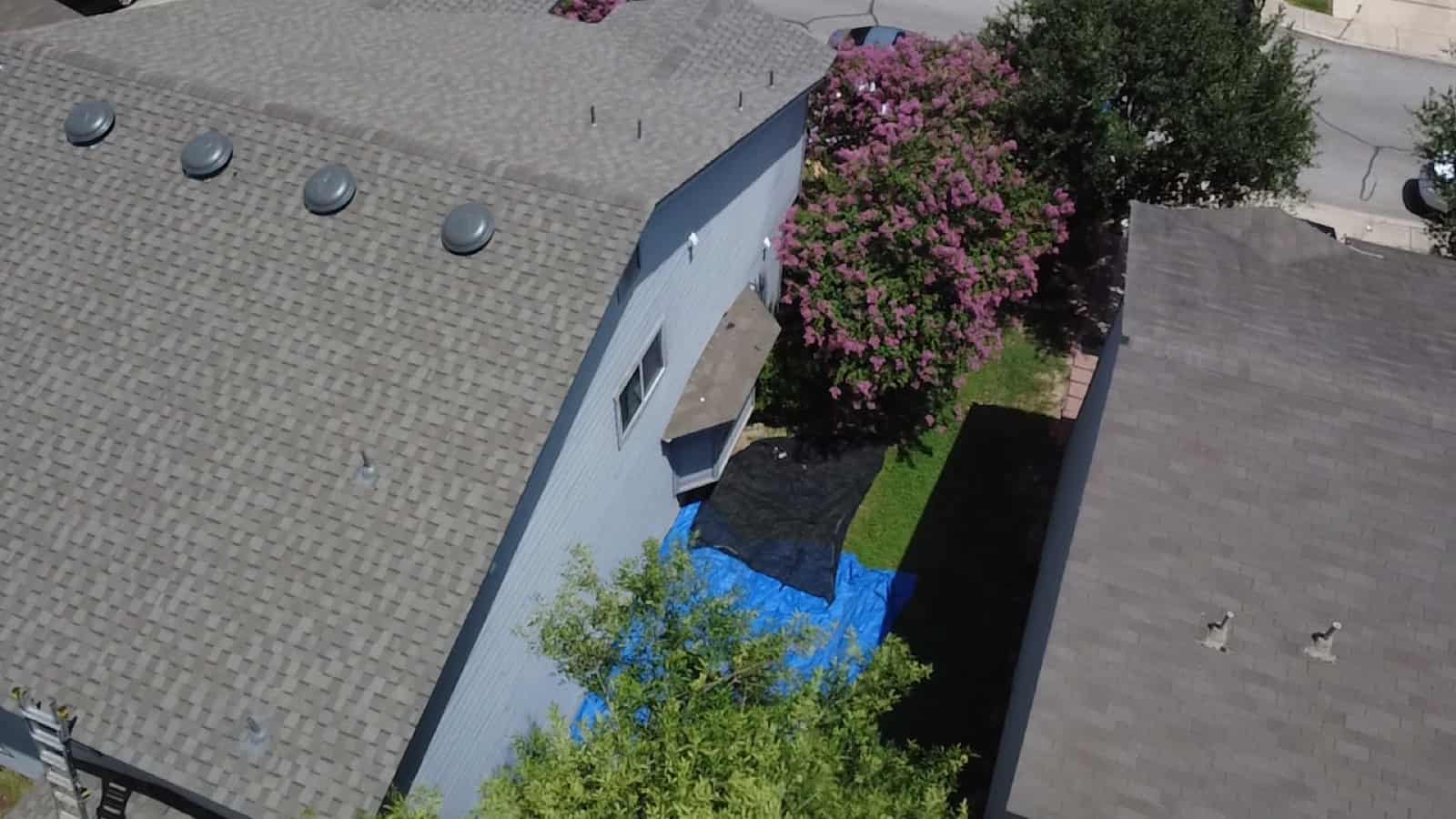 Aerial view of a San Antonio shingle roof with ridge vents and an emergency tarp staged in the yard after a leak