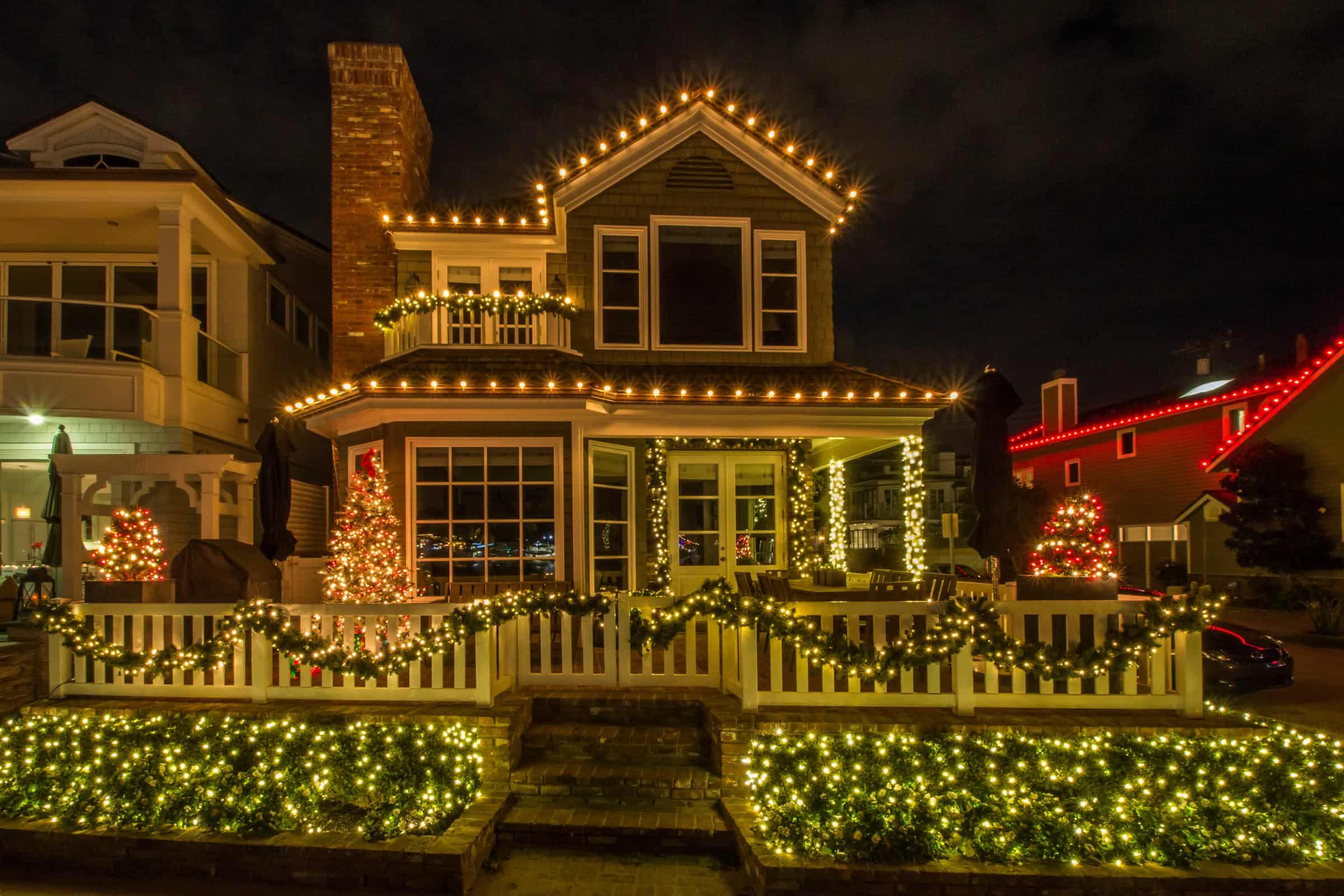 Two-story home at night with warm white bulbs evenly spaced along every roofline, eave and gable, lit garland on the porch and picket fence, and net lighting across the front hedges